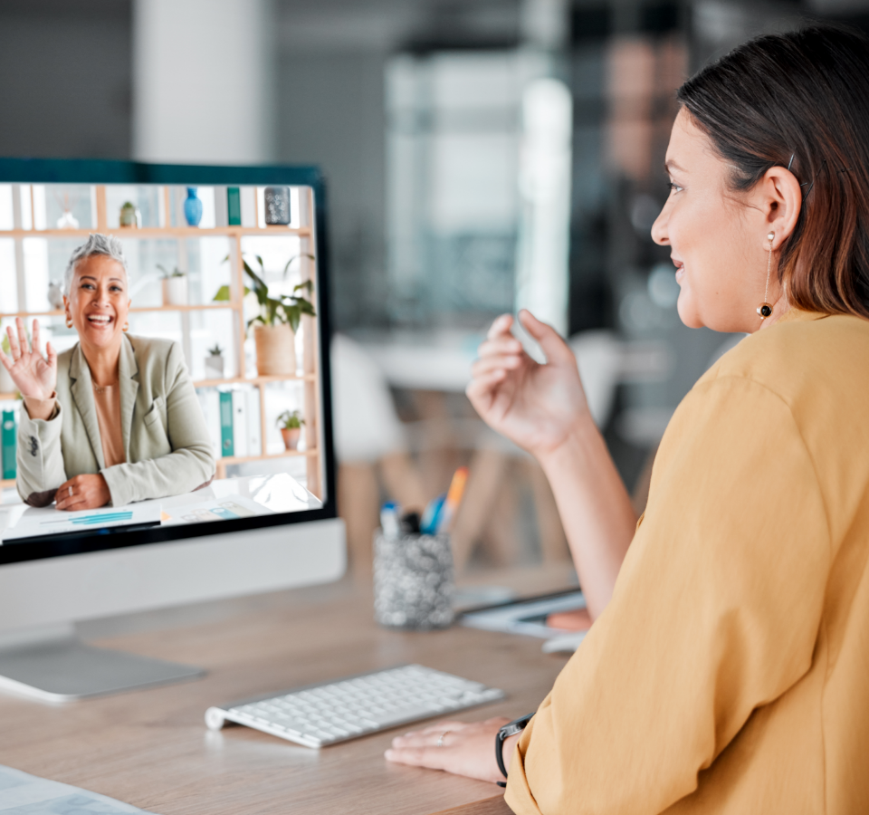 A woman sits in front of a screen and greets another woman who is connected to her via video telephony.